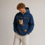 A man wearing a blue denim-style hooded jacket with a cat on the front pocket, paired with light beige pants, against a plain light gray studio background.