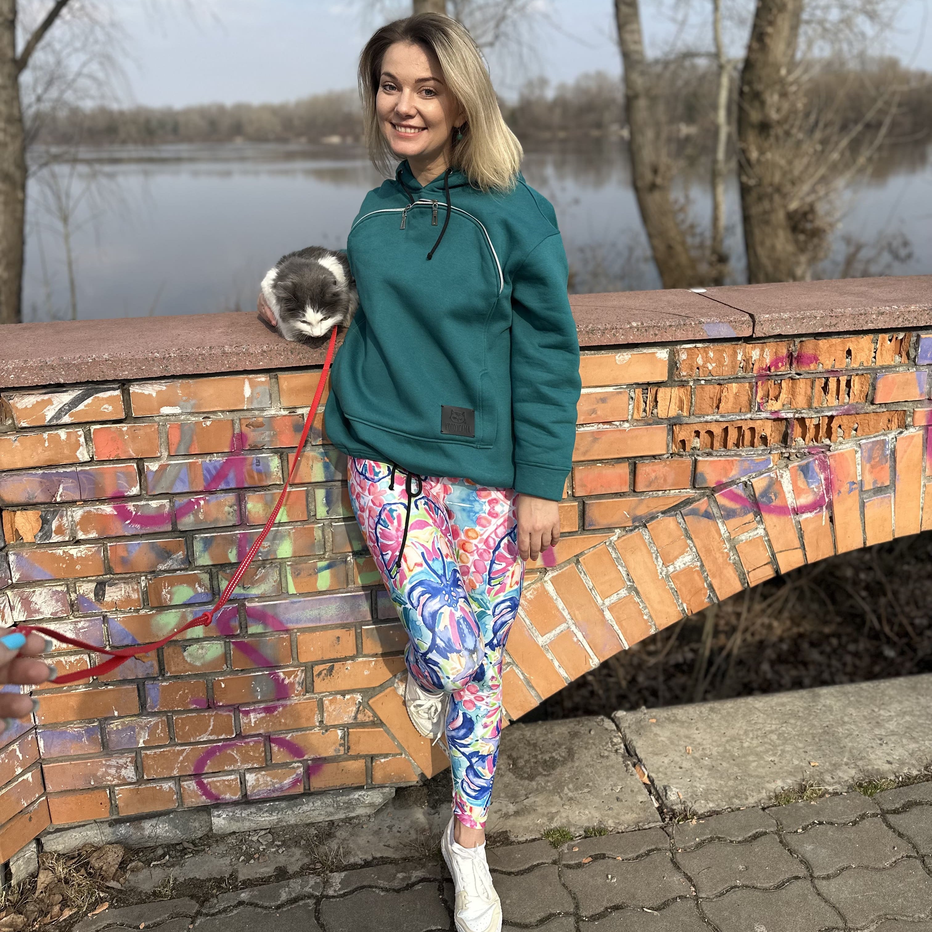 Woman in teal jacket and colorful leggings standing by a brick wall with a lake in the background