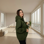 A woman wearing an oversized dark green pet hoodie with a small dog in a carrier pocket, standing in a bright, modern room with large windows.