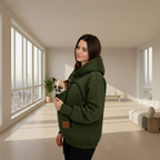 A woman in a modern room wearing an dark green hooded pet carrier sweatshirt with a small dog in the front pocket, looking over her shoulder.
