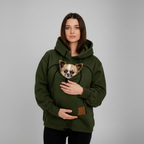 A woman wearing an dark green hoodie with a built-in pet pocket holding a small Chihuahua dog, photographed against a neutral gray studio background.