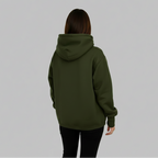 A person viewed from behind wearing an dark green pullover hoodie and black pants, standing in a bright, modern minimalist room with large windows and natural light.