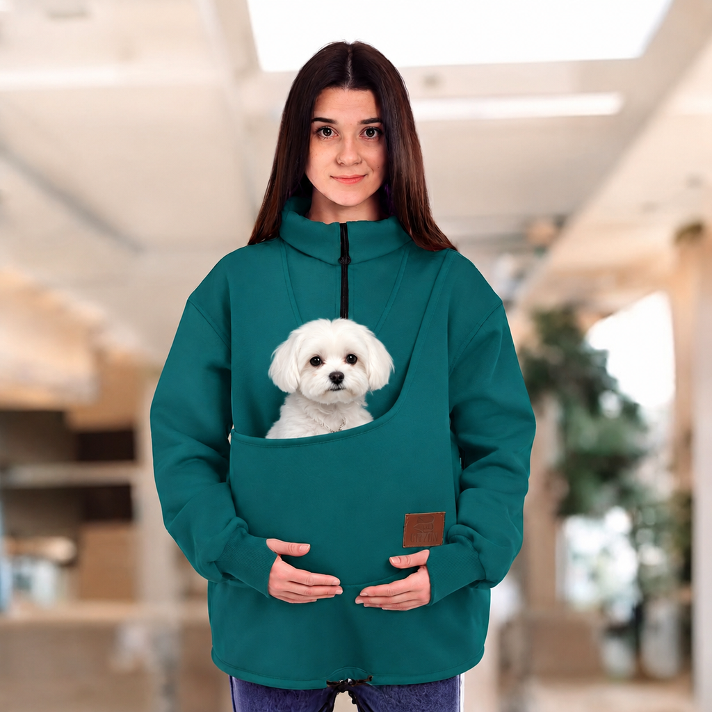 Women wearing a teal sweatshirt with a maltipoo dog in pouch, standing indoors.