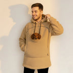 Man wearing a beige boucle hoodie with a pocket containing a small dog, standing against a plain background.