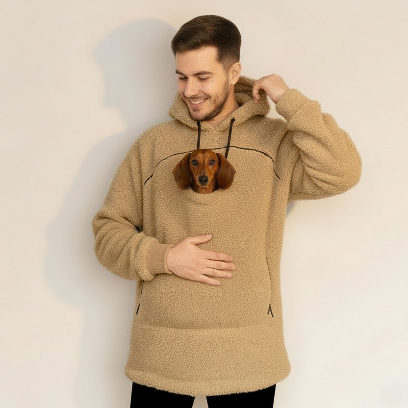 Man wearing a beige boucle hoodie with a pocket containing a small dog, standing against a plain background.