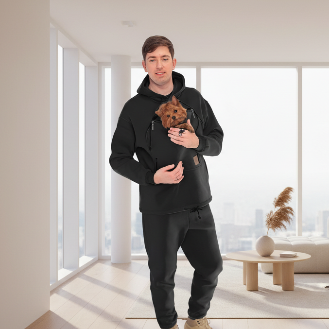 Man holding a small dog in a black hoodie with a pocket, standing in a modern living room.