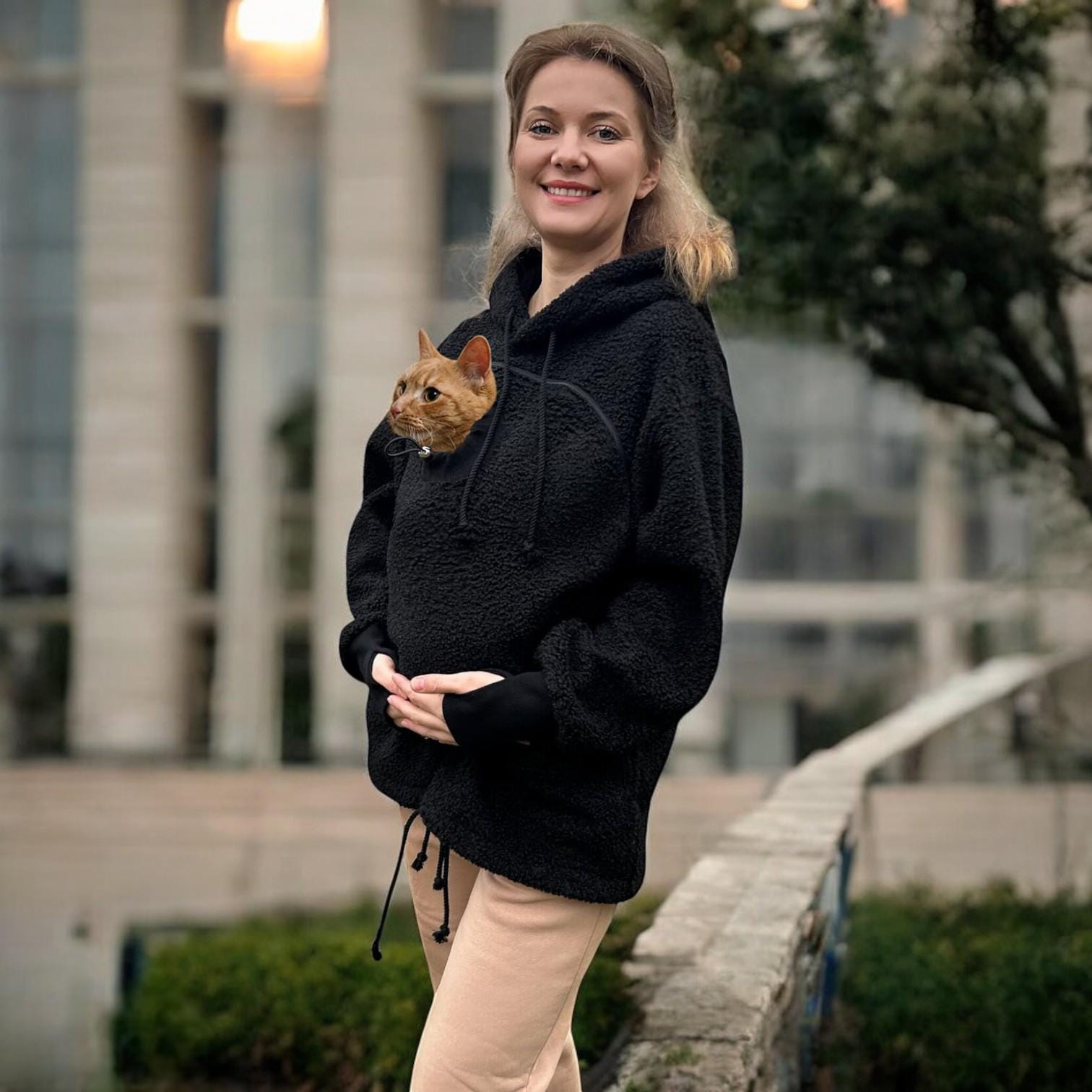 Woman wearing a black boucle hoodie with a cat peeking out, standing outdoors.