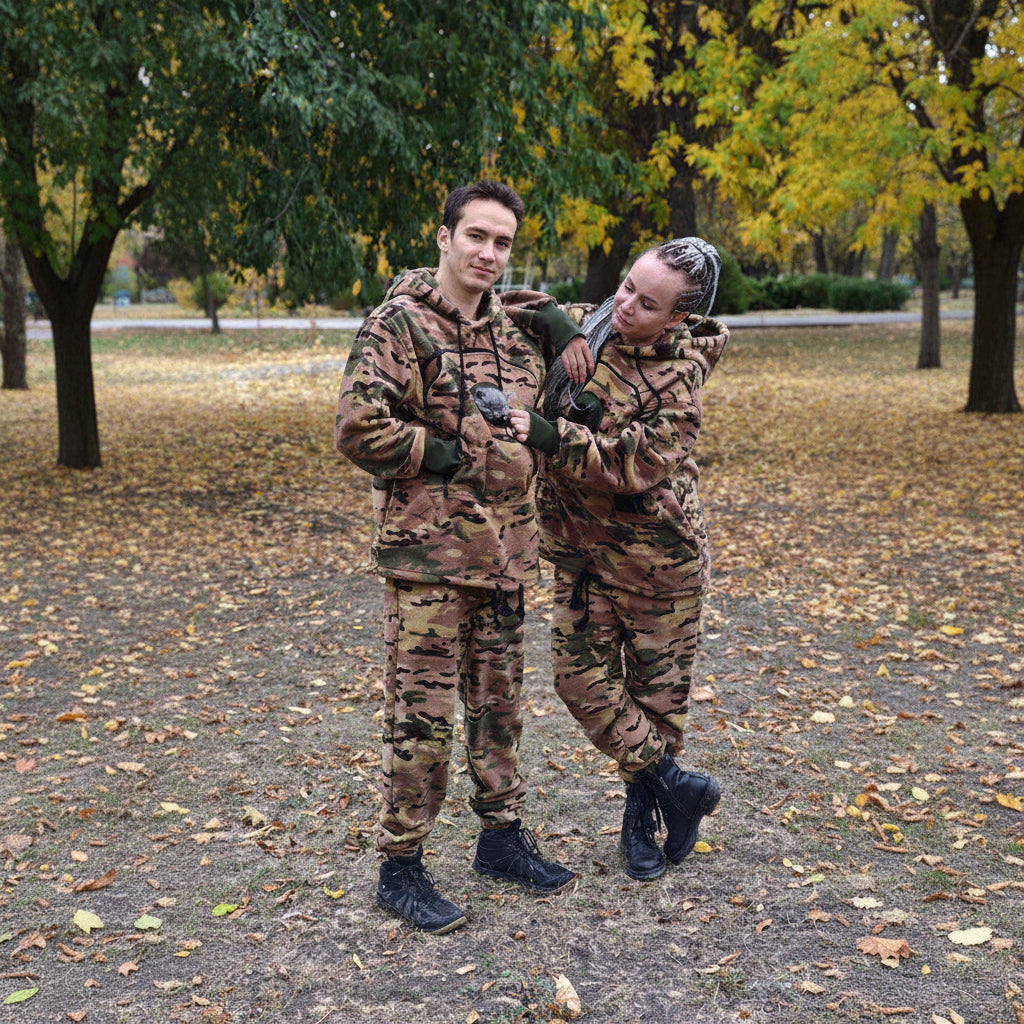 Two people in camouflage outfits standing outdoors with trees and fallen leaves in the background.