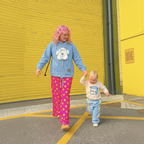 A woman with pink hair and a toddler walking hand in hand in front of a bright yellow corrugated metal building. The woman wears a light blue hoodie with a white 3 d flower graphic and bright pink pants with yellow polka dots.