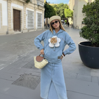 A woman stands on a city street wearing a light blue matching lounge set with a flower applique featuring an orange cat on the chest. She is also wearing a beige hat, sunglasses, and carrying a woven straw handbag.