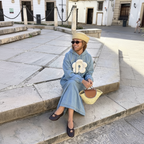 A woman sits on stone steps in an outdoor plaza wearing a light blue denim hoodie with a large cream flower applique on the chest, a beige beret, dark sunglasses, and dark loafers. She holds a woven straw handbag with a brown leather detail.