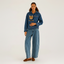A model stands against a light gray studio background wearing a blue denim hoodie with a cat in the front pocket, matching wide-leg denim jeans, and brown suede shoes.
