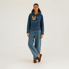 A model stands against a light gray studio background wearing a blue denim hoodie with a cat in the chest, matching wide-leg denim jeans, and brown suede loafers.