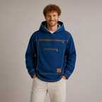 A smiling man with curly red hair wearing a blue hooded jacket with a front zip pocket and white pants, standing against a plain light gray studio background.