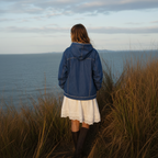 A woman viewed from behind wearing a blue denim hooded jacket over a white lace-trim skirt, standing in tall grass looking out toward a body of water under a cloudy sky.