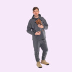Man wearing a gray hoodie and pants holding a small dog, standing against a white background