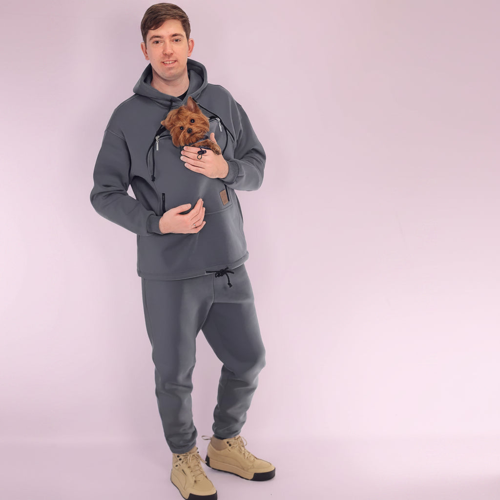 Man wearing a gray hoodie and pants holding a small dog, standing against a white background Main image