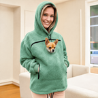 Person wearing a green hoodie with a dog peeking out of a pocket, sitting on a couch.