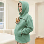 Person wearing a green hoodie with a small dog peeking out of the pocket, standing in a room.