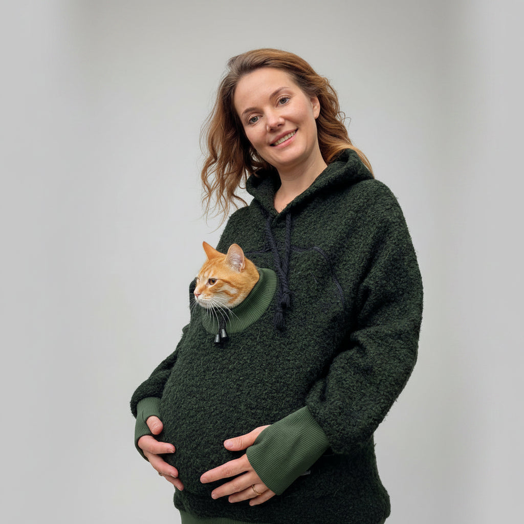 A woman wearing a dark green fuzzy hoodie with a cat pocket, holding an orange tabby cat in the pocket, set against a grey background