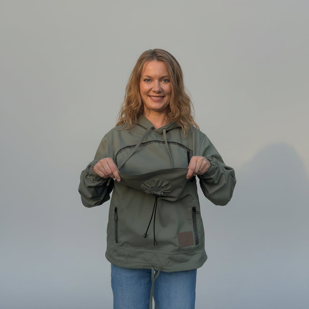A woman wearing an olive green hooded jacket with a drawstring pouch on the chest, holding it open to show the interior. She is wearing blue jeans and standing in front of a beige fabric backdrop.