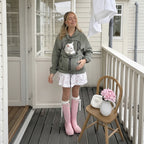 A woman stands on a porch wearing an olive green hooded sweatshirt with a front pet carrier pocket containing a white cat. She is also wearing a white ruffled skirt, pink rain boots, and holding a small white purse.