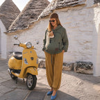 A pregnant woman wearing an olive green hoodie with a cat carrier, mustard yellow pleated pants, and blue shoes stands next to a yellow Vespa scooter in front of white stucco domes.