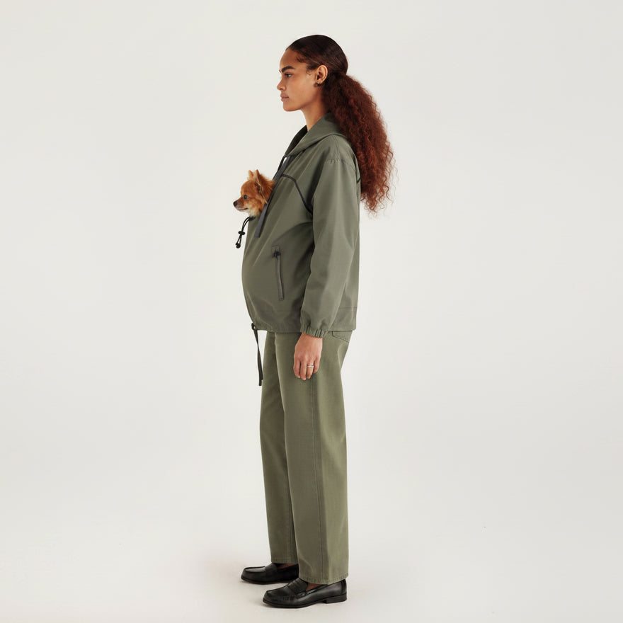 A woman in profile wearing an olive green utility jacket and matching wide-leg trousers, holding a small brown dog against her chest. She is standing against a plain white studio background.