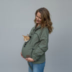 A pregnant woman wearing an olive green cat carrier jacket with an orange tabby cat peeking out from the front pouch, standing against a beige wrinkled backdrop.