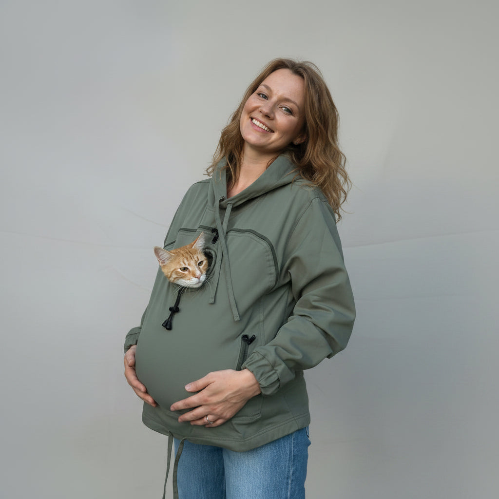 A pregnant woman wearing an olive green hoodie with a cat peeking out of the chest pocket, standing against a wrinkled beige backdrop.