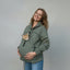 A pregnant woman wearing an olive green hoodie with a cat peeking out of the chest pocket, standing against a wrinkled beige backdrop.