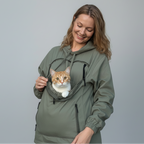 A woman wearing an olive green hooded jacket with a front cat pocket, smiling as an orange and white cat peers out from the pocket, against a solid gray studio background.