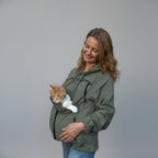 A woman wearing an olive green hooded jacket with a cat carrier pocket, holding an orange and white cat against her stomach, set against a wrinkled beige backdrop.
