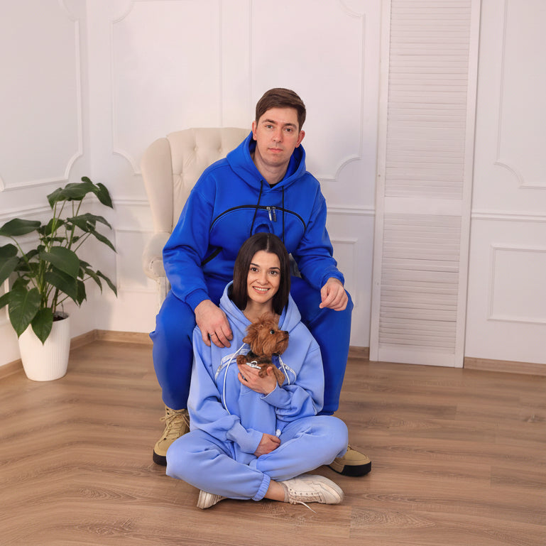 Two people in matching blue and light blue onesies sitting on the floor with a small dog.