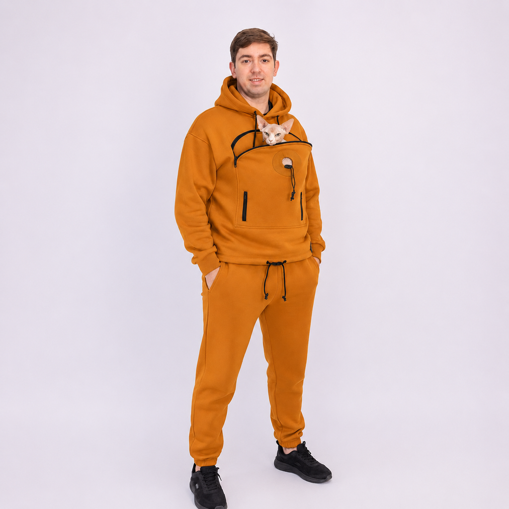 Man wearing a mustard yellow tracksuit with a small cat peeking out of the front pocket on a white background