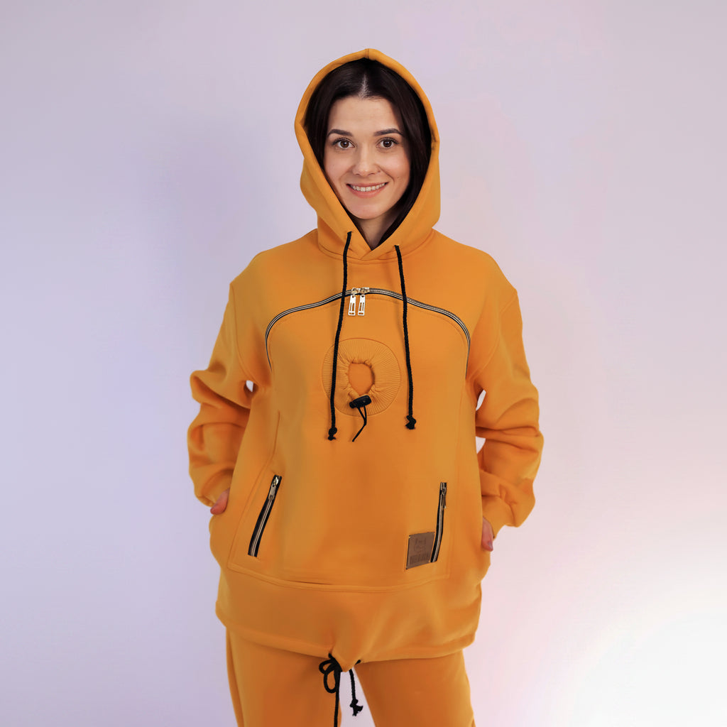 Person wearing a mustard hoodie with zippers on a light purple background