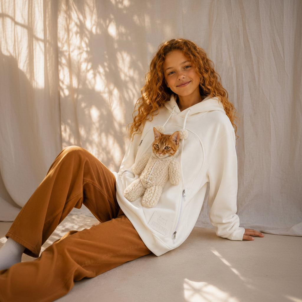 A smiling young girl with long curly red hair sits on a floor wearing a white zip-up hoodie and brown pants, holding a small orange tabby kitten wrapped in a knit blanket. The background is a draped cream fabric with dappled sunlight and shadows.