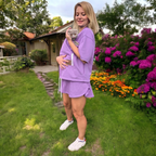 Woman in a purple outfit holding a cat in a garden with flowers and a house in the background