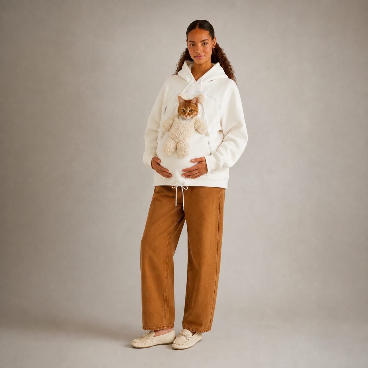 A woman stands against a plain light gray studio background wearing a white hoodie with a cat stuffed inside the front pocket, paired with wide-leg brown pants and cream slip-on shoes. Main image
