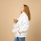 A woman wearing a white fleece hoodie with a built-in plush cat pocket, holding an orange tabby cat inside. She is standing against a solid beige studio background.