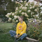 A woman sits cross-legged on a garden bench in front of a large bush of white flowers, wearing a bright yellow hoodie with a pink flower applique, blue jeans, and green gardening boots while holding a small white cup.