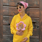 A woman wearing a bright yellow hoodie, pink striped beanie, and round sunglasses holds a ginger cat wearing a pink flower-shaped pet collar against a rustic wooden door background.