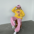 A person wearing a bright yellow hoodie with a pink flower graphic containing a cat, paired with pink sweatpants and a pink bucket hat, sitting on a wooden stool in a minimalist room.