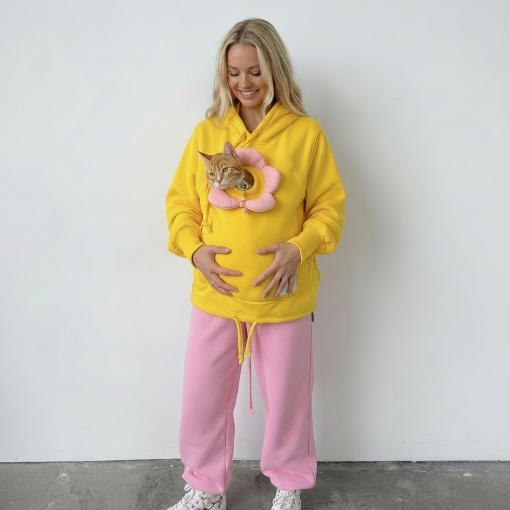 A woman wearing a bright yellow hoodie with a pink flower-shaped belly ornament and a cat nestled inside, paired with pink sweatpants, standing against a plain white wall.